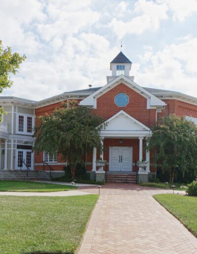 A brick and white-trimmed building with a central cupola and a curved pathway leading to the entrance, flanked by green trees and a signpost in the foreground.